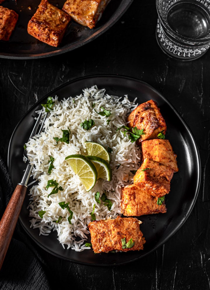 A balck plate with tandoori salmon on the right side and rice on the left side with a brown handled fork.