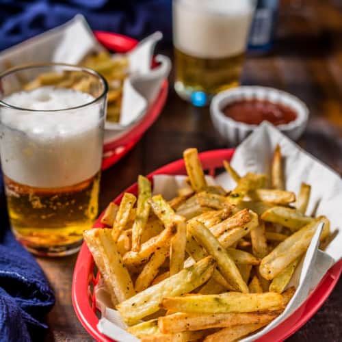 A red basket of air fryer french fries in the front with a blue towel, two glasses of beer, a small bowl of ketchup, and another red basket of french fries in the back on a wooden table.