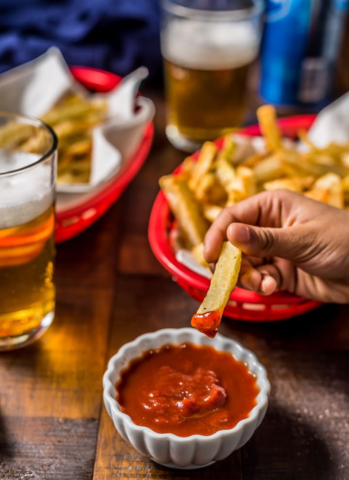 A hand dipping an air fryer french fry into a small bowl of ketchup with two red baskets filled with fries and two glasses of beer on a wooden table.