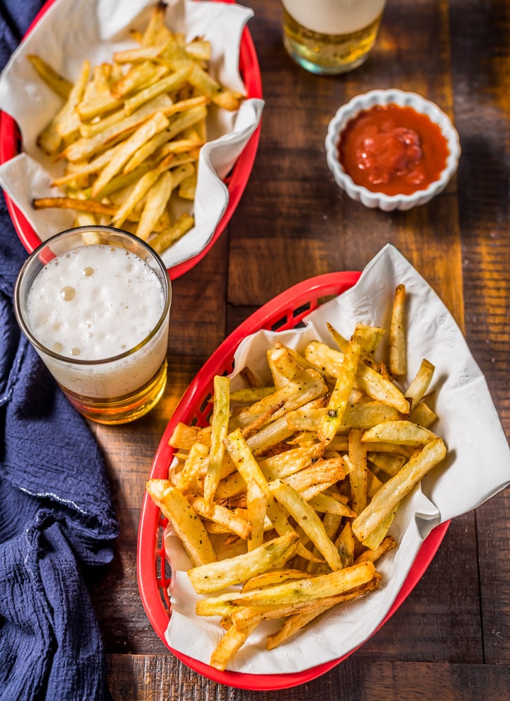 A picture from the top of two red baskets of french fries with a blue towel, a small bowl of ketchup, and two beers on a wooden table.
