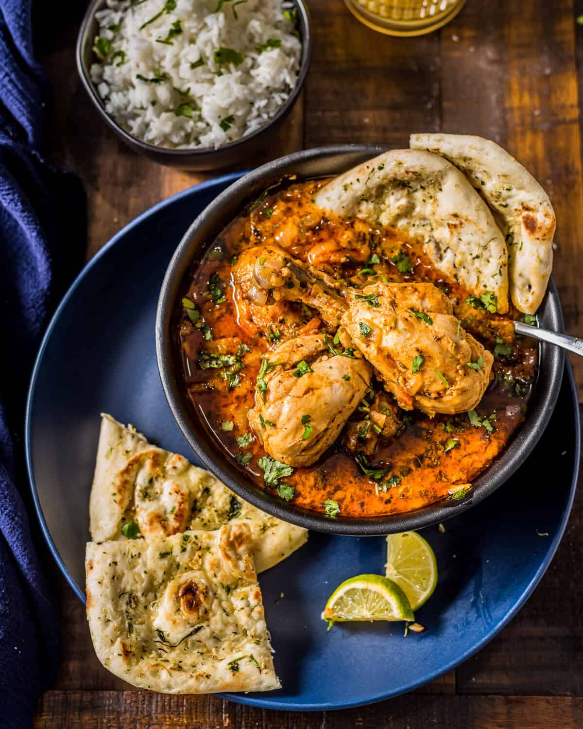 An overhead shot of Indian chicken curry served with naan, rice, lemon wedges and Mango lassi