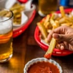 A hand dipping a french fry into a bowl of ketchup with red baskets of french fries in the back and two beers on a wooden table and the words Perfect Air Fryer French Fries at the top.