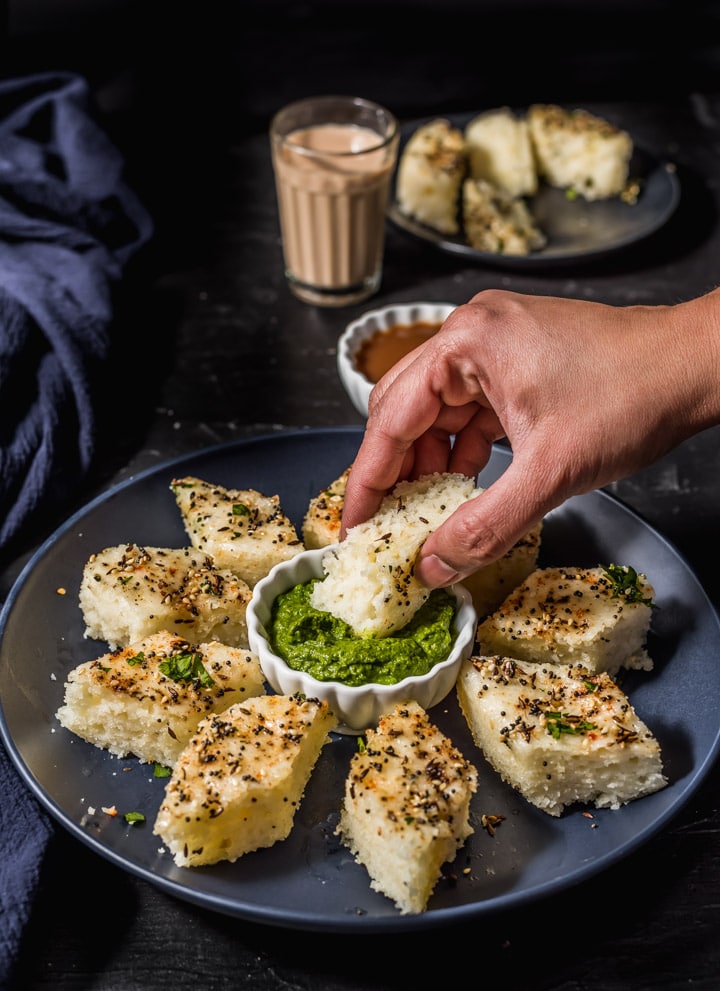 A hand dipping a piece of rava dhokla in chutney.