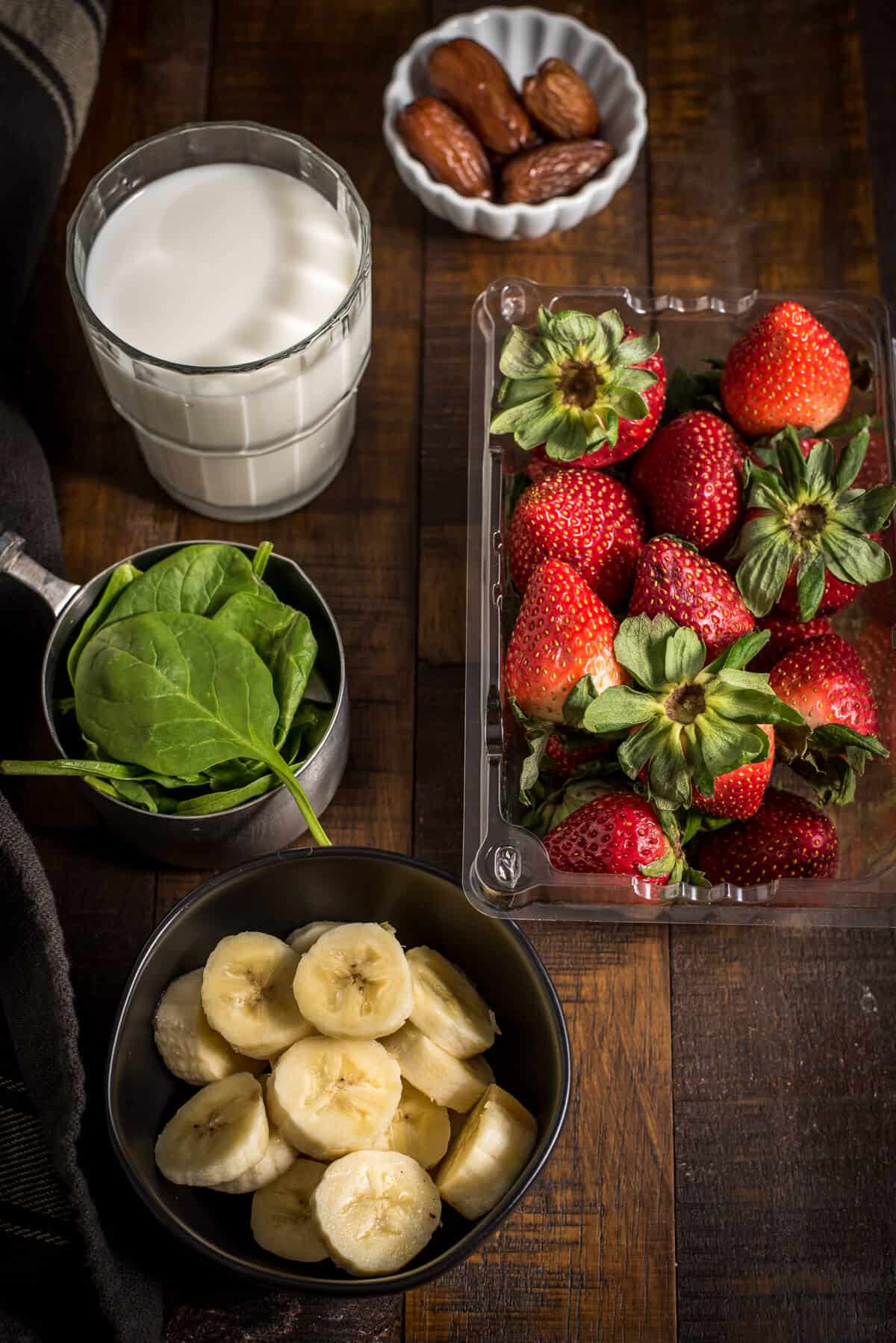 Strawberry Banana Spinach Smoothie Ingredients such as spinach, milk, banana, strawberry and dates are kept on a wooden surface