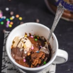 A white coffee mug with a chocolate mug cake inside topped with vanilla chocolate ice cream, sprinkles and chocolate sauce with a fork sticking out of the mug.