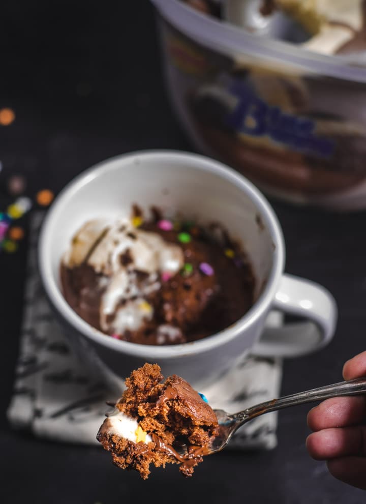 A white mug with chocolate mug cake and melted vanilla and chocolate ice cream on top with a fork full taken out. A fork in front of the mug shows a bite of the chocolate mug cake.