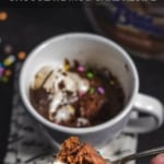 A white mug with chocolate mug cake and melted vanilla and chocolate ice cream on top with a fork full taken out. A fork in front of the mug shows a bite of the chocolate mug cake. The words Chocolate Mug Cake Recipe are at the top of the photo.