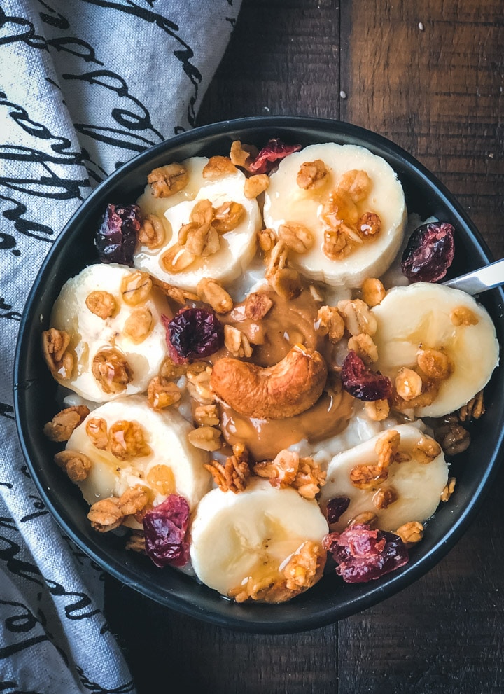 A black bowl with instant pot oatmeal topped with banana and oats on a wooden table with a dish towel to the left.