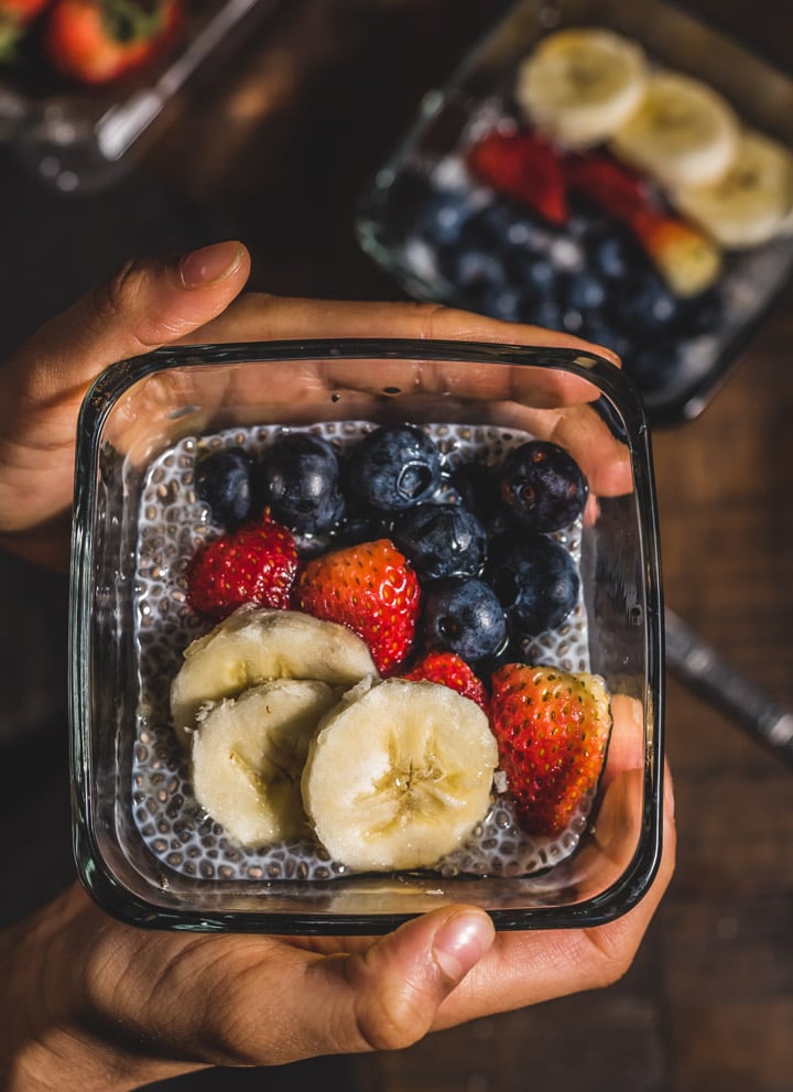 A glass blow with chia pudding topped with banana, strawberry, and blueberry in two hands.