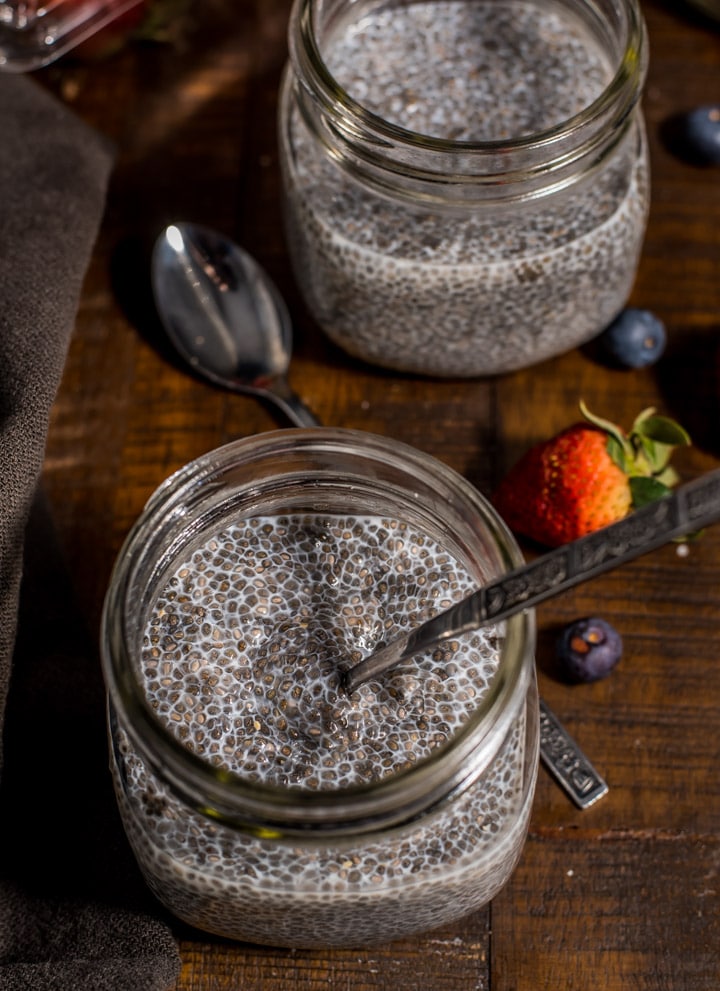 A mason jar with chia seed pudding with a spoon in it and another jar of chia seed pudding in the back on a wooden counter with blueberries and a strawberry and an additional spoon on the counter.
