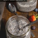 A mason jar with chia seed pudding with a spoon in it and another jar of chia seed pudding in the back on a wooden counter with blueberries and a strawberry and an additional spoon on the counter with the words Healthy Chia Pudding Recipe at the top.