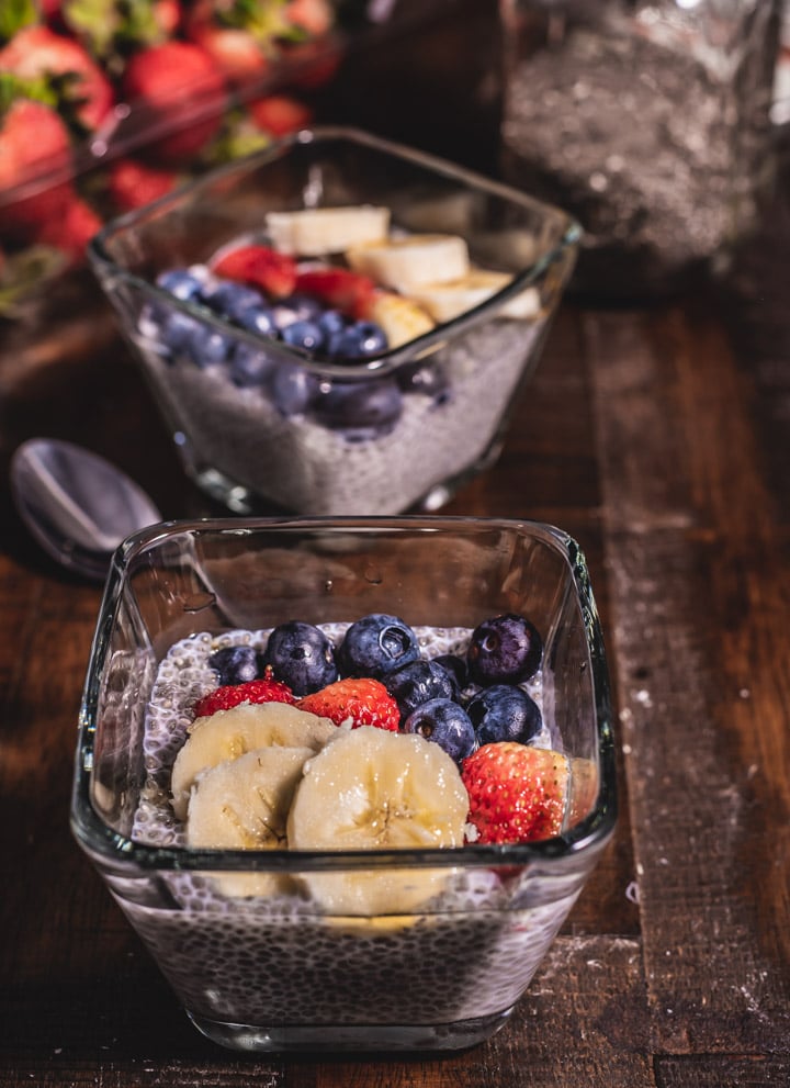 Two glass bowls of vanilla chia pudding topped with banana, strawberry, and blueberry on a wooden table with a spoon in between them and strawberries behind them.