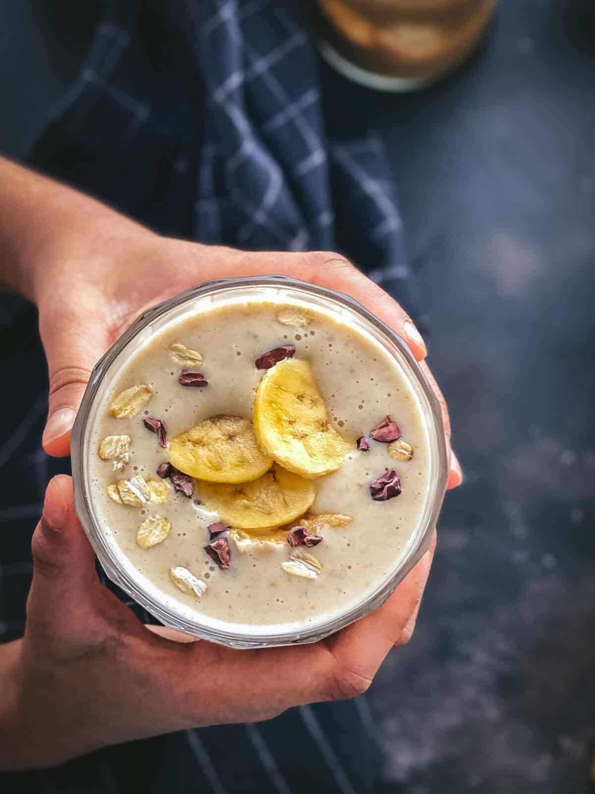 A hand holding a glass of Peanut Butter Banana Smoothie topped with banana and oats