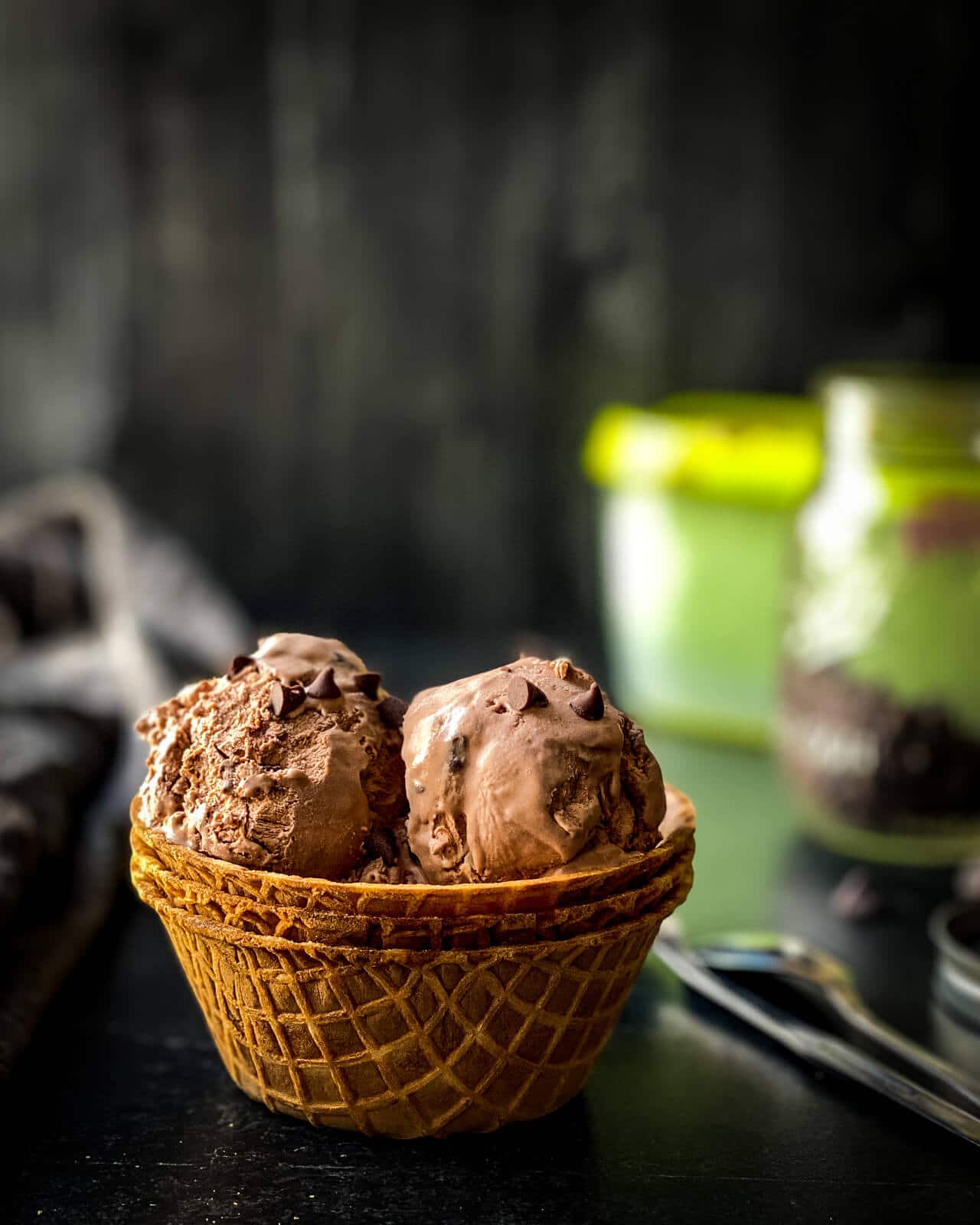 A waffle cone bowl with scoops of double chocolate ice cream on a wooden counter with a container of ice cream in the back.