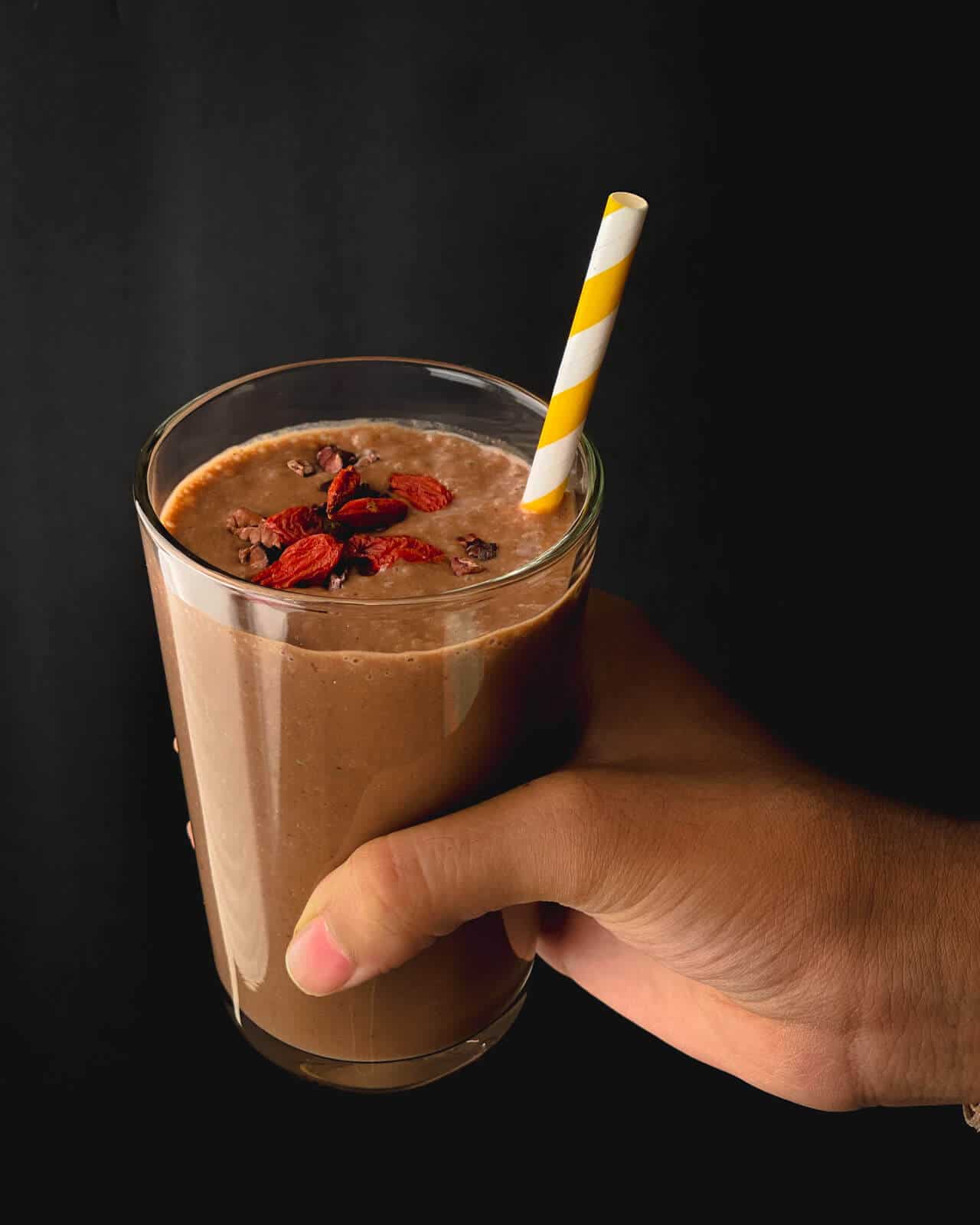 A hand holding a banana chocolate smoothie with goji berries on top and a yellow and white straw.