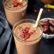 A smoothie on a black counter with goji berries and a yellow and white straw on top. Behind the smoothie is a bowl of dates, a banana, and another chocolate banana smoothie.