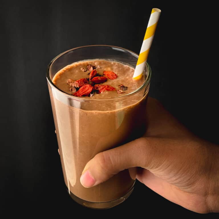 A hand holding a chocolate date smoothie in a clear glass cup with goji berries on the top of the smoothie and a yellow and white straw in the smoothie.