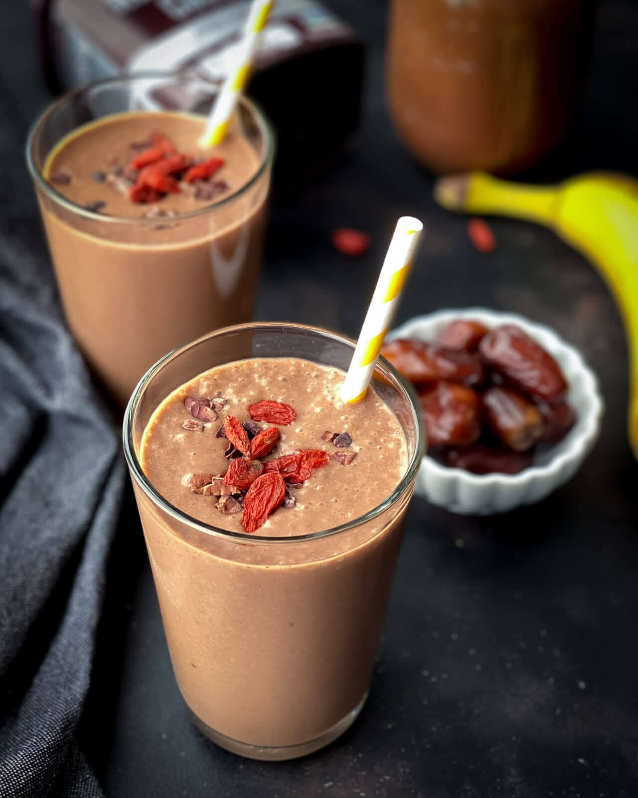 A chocolate date smoothie in the front with goji berries on top. A small white bowl of dates behind the front smoothie with a banana and an additional smoothie to the left on a black counter.