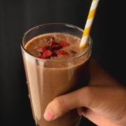 A hand holding a clear glass filled with chocolate smoothie, topped with goji berries, and a white and yellow straw sticking out with the words Chocolate Smoothie at the top in yellow.