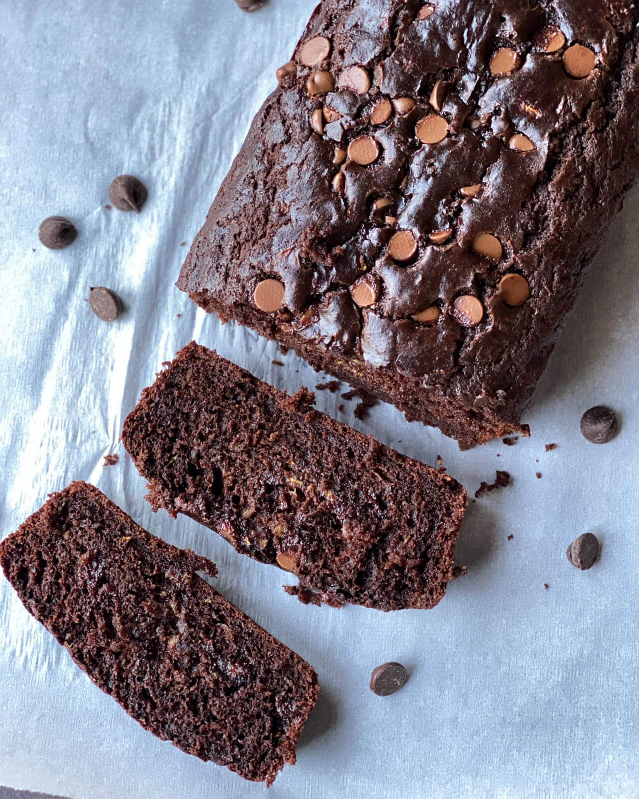 A loaf of double chocolate zucchini bread with two slices laying in front on a sheet of parchment paper.