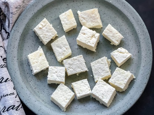 Cut cubes of paneer on a light blue speckled plate with a dish towel to the left.