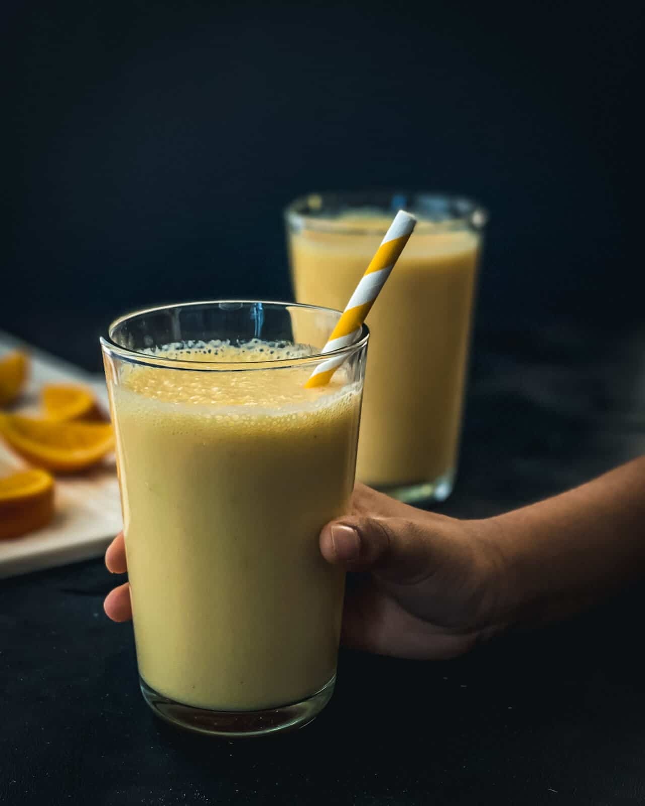 Two glasses of orange smoothie on a counter with a hand holding the front glass with a white and yellow straw.