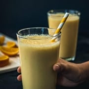 Two glasses of orange smoothie on a counter with a hand holding the front glass with a white and yellow straw and the words Orange Julius Smoothie at the top.