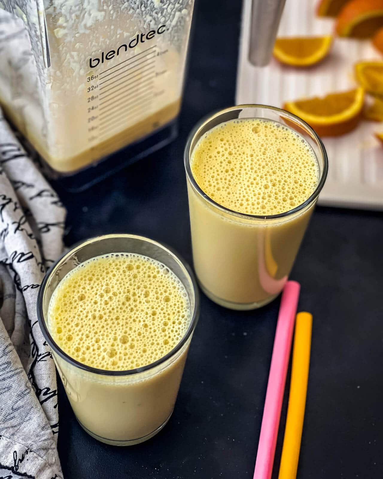 Two glasses of orange julius on a counter with two straws to the right, a cutting board of orange slices in the back, and a blender with orange smoothie in the back left.