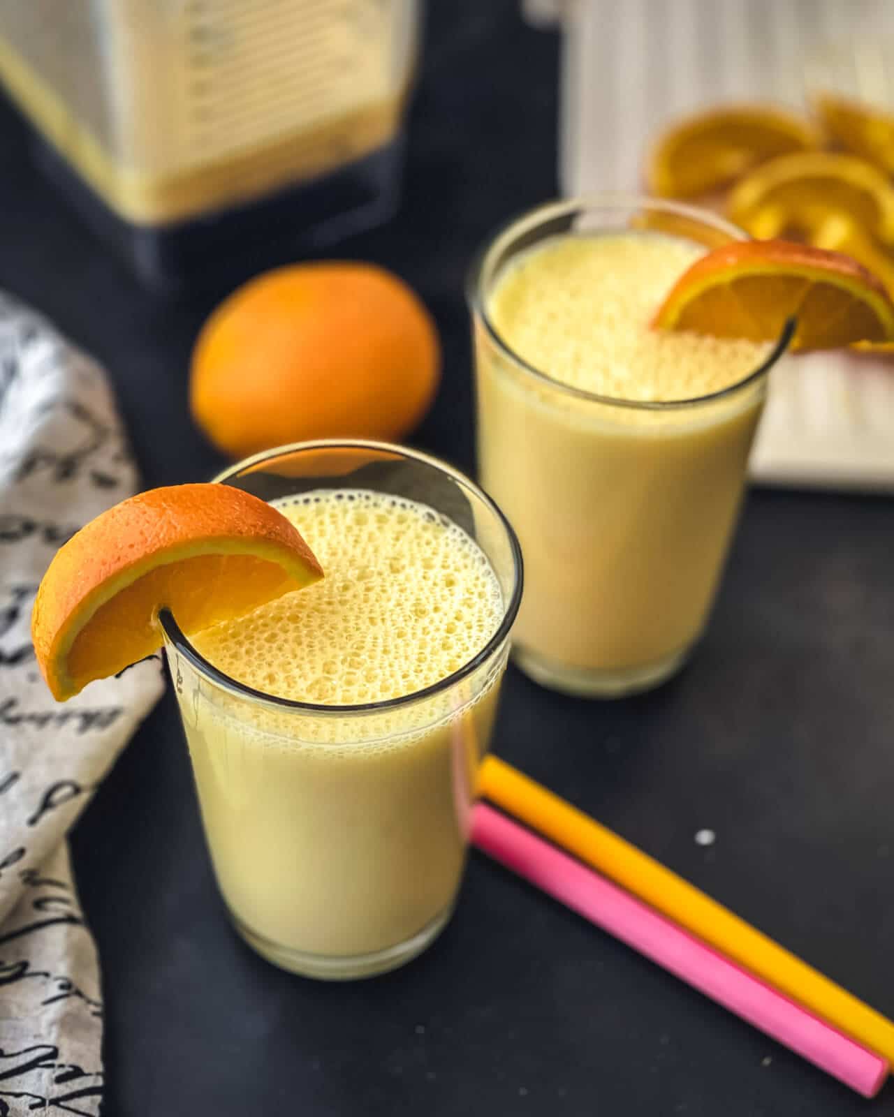Two glasses of orange smoothie with an orange slice on the rim, two straws in between, and an orange in the back.