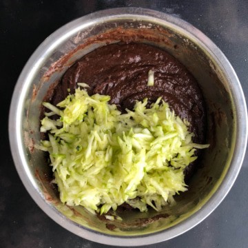 A silver mixing bowl with the zucchini shreds on top of the chocolate bread batter.