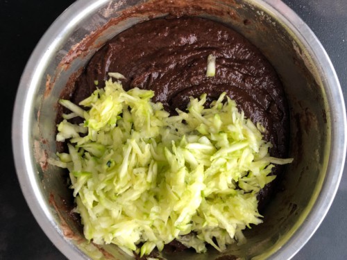 A silver mixing bowl with the zucchini shreds on top of the chocolate bread batter.