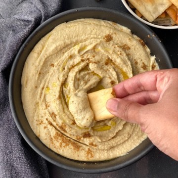 A hand dipping a pita chip into a black bowl filled with creamy instant pot hummus.