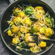 A black bowl with cheese topped steamed Brussel sprouts with the words Cheesy Brussel Sprouts at the bottom in yellow and a silver fork in the bowl.