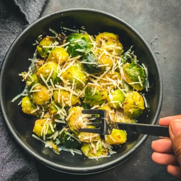 A black bowl of cheese topped steamed and sautéed Brussel sprouts with a silver fork poking into a Brussel sprout on a grey counter.