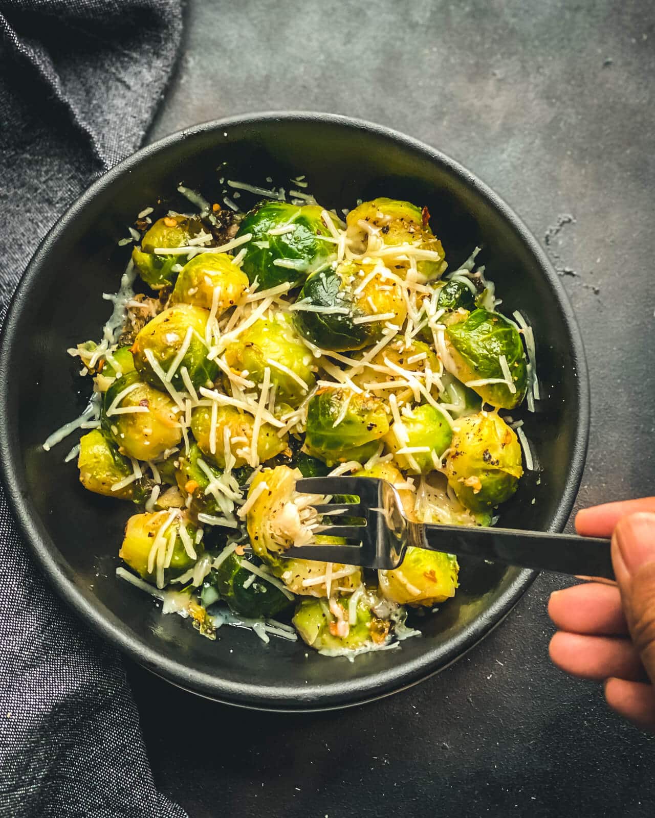 A black bowl of cheese topped steamed and sautéed Brussel sprouts with a silver fork poking into a Brussel sprout on a grey counter.