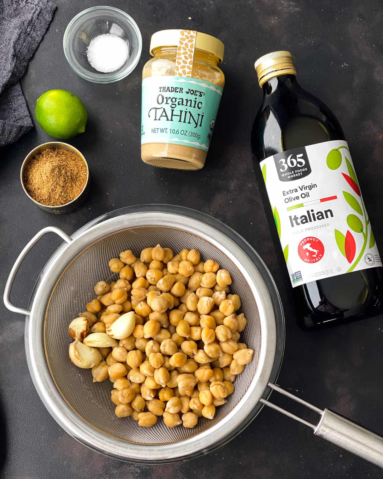 Chickpeas in a strainer with a bottle of olive oil, a jar of tahini, salt, a lime, and cumin above.