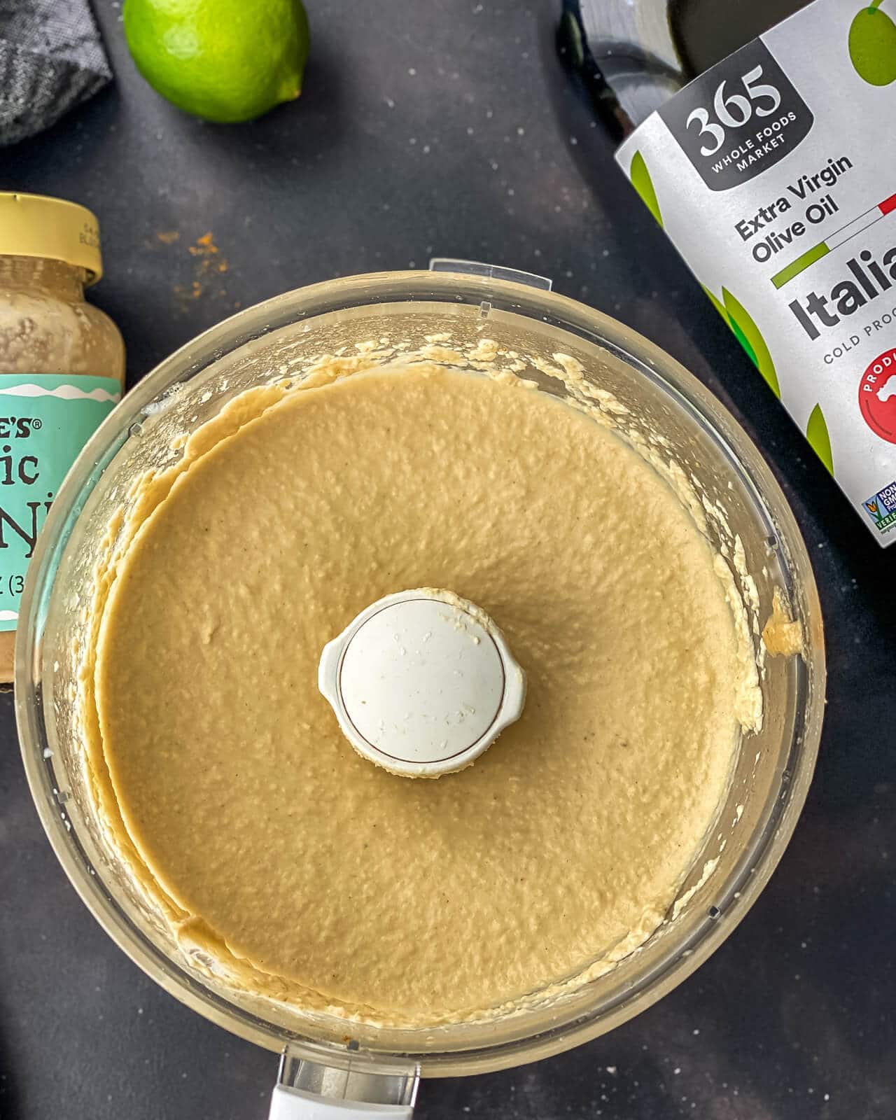The base of a food processor with creamy blended hummus, a bottle of olive oil, a lime, and a jar of tahini around the hummus.