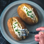 The words Instant Pot in the top right in white with the words baked potato below in yellow and a blue plate in the center with two baked potatoes topped with sour cream and chive and a hand holding a fork poking into one of the potatoes.