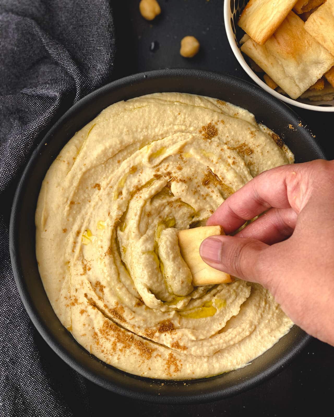 A hand with a pita chip dunked into the bowl of hummus with a small bowl of pita chips to the top right.
