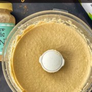 The base of a food processor with creamy blended hummus, a bottle of olive oil, a lime, and a jar of tahini around the hummus with the words Easy Hummus Recipe at the top.