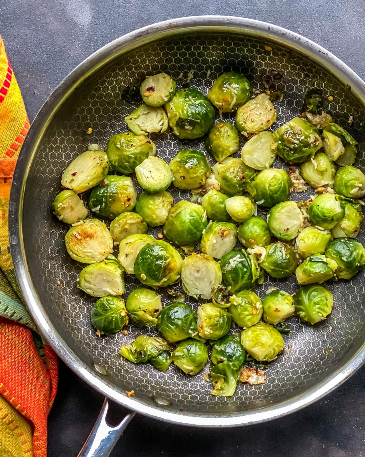 A silver pan of pan fried steamed Brussel sprouts on a grey counter with an orange and yellow towel to the left.