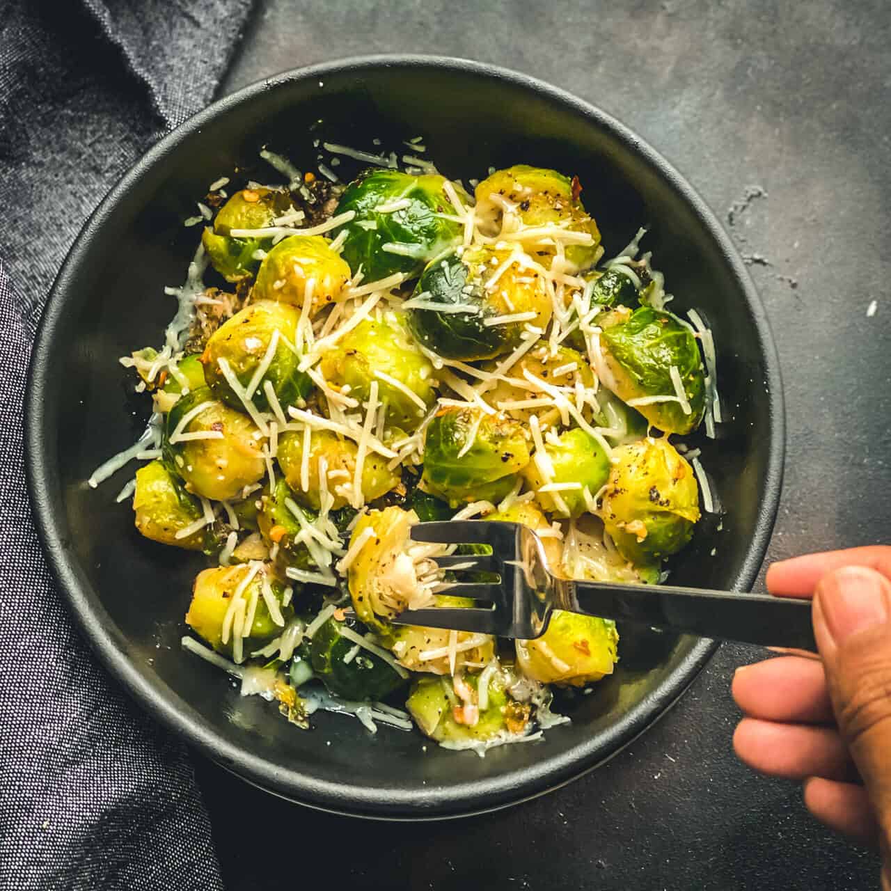 A black bowl with steamed and sautéed Brussel sprouts topped with cheese and a silver fork being poked into the bowl on a grey counter.