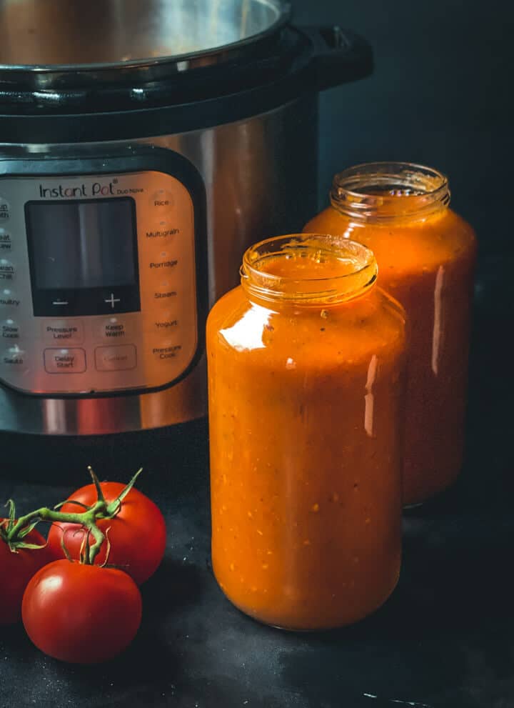 Two jars of marinara sauce in front of the instant pot and two tomatoes on the vine.