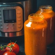Two glass jars of pasta sauce in front of an instant pot with two tomatoes on the vine to the left with the words Instant Pot Classic Marinara Sauce at the top.