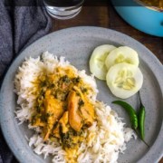 A light grey plate with a scoop of rice topped with methi chicken and a side of cucumbers on a wooden table with a grey towel to the left and the words Instant Pot Fenugreek Chicken at the top.