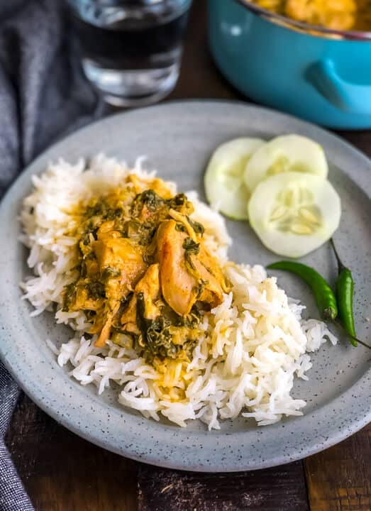 A light grey plate of methi murgh on rice with three cucumbers to the right on a wooden counter.