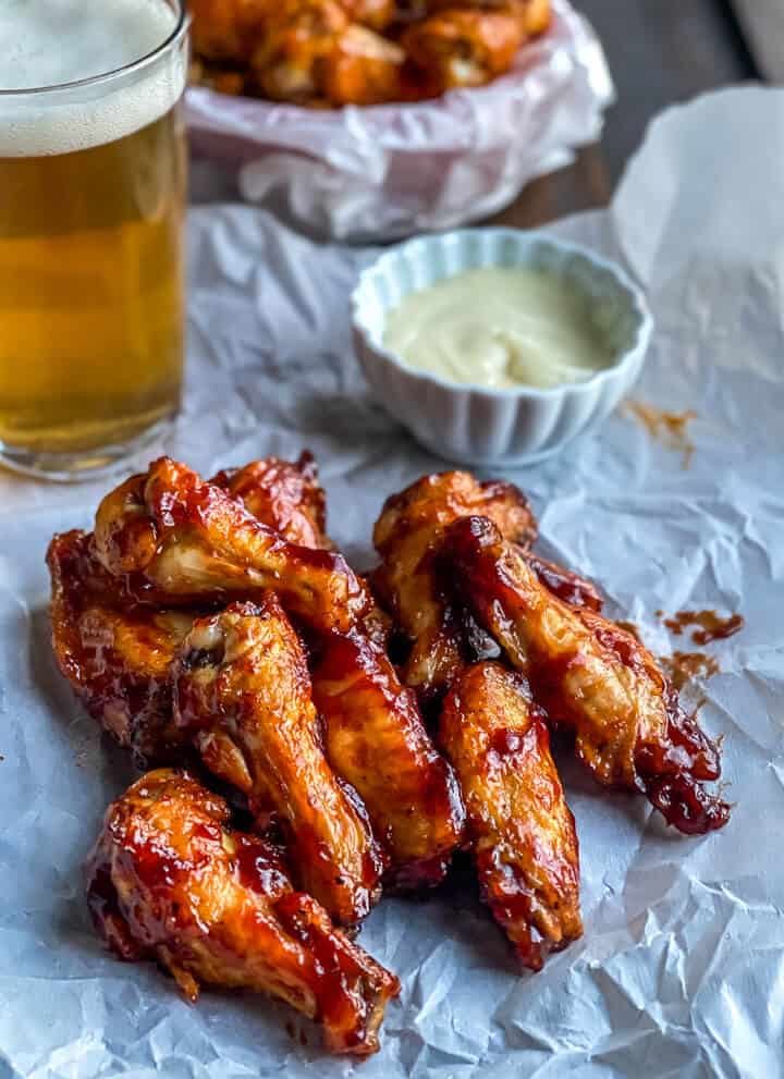 A stack of chicken wings coated in bbq sauce with a bowl of ranch in the back and a cup of beer to the left.
