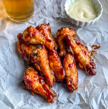 A parchment paper with a stack of chicken wings with a small bowl of ranch to the right and a cold beer in the back left.