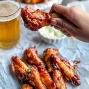 A hand holding a bbq chicken wing with other wings on a piece of parchment paper below and the words Air Fryer BBQ Chicken Wings at the top.