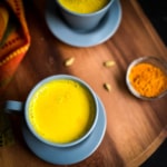 A blue mug with golden turmeric latte on a wooden counter with another latte in the back and a small bowl of turmeric to the right.